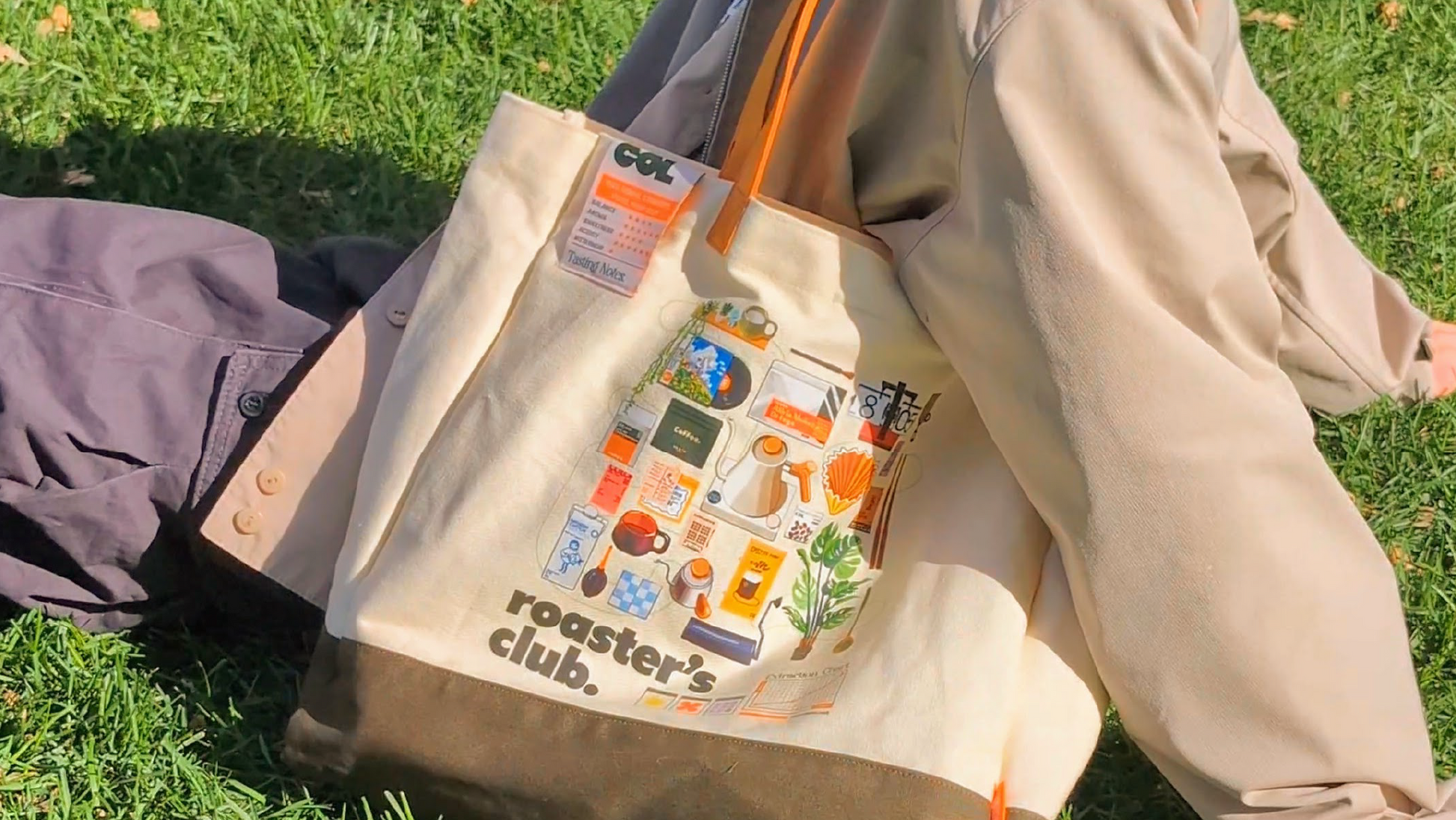 Person sitting on grass with a beige tote bag featuring 'roaster's club' branding.